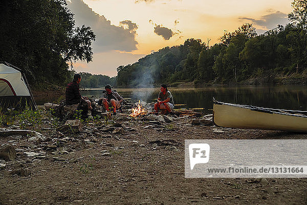 Freunde unterhalten sich  während sie bei Sonnenuntergang am Campingplatz gegen den Himmel sitzen