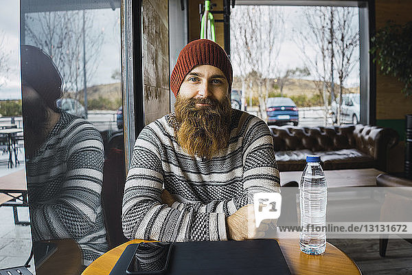 Portrait of confident hipster man with smart phone and laptop computer on table at cafe