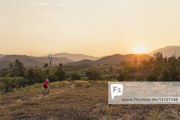 Hochwinkelansicht eines Mannes beim Mountainbiking auf dem Feld gegen den Himmel bei Sonnenuntergang