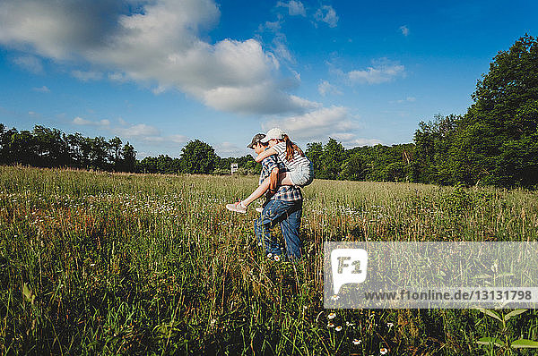 Seitenansicht des Bruders  der seine Schwester Huckepack nimmt  während er im Park auf einem Grasfeld gegen den blauen Himmel läuft