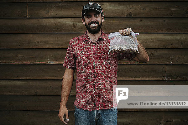Portrait of man holding peanuts in plastic bag while standing against wooden wall