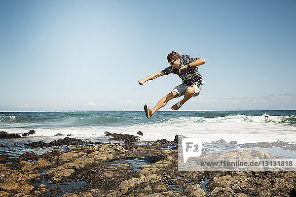 Man jumping on rocky shore against blue sky