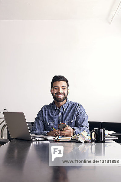 Smiling businessman using phone while sitting at table against wall in office