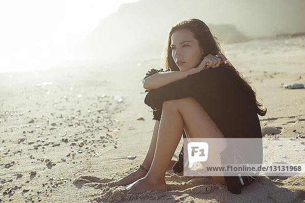 Nachdenkliche Frau umarmt die Knie  während sie am sonnigen Tag am Strand sitzt