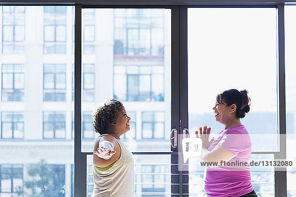 Happy friends exercising face to face while standing against window in yoga studio