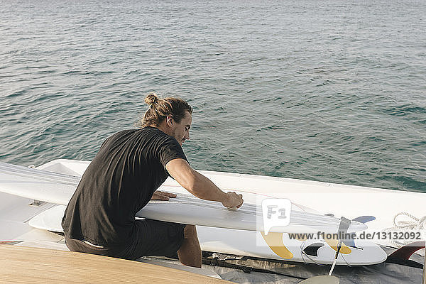 Rear view of man cleaning surfboard while sitting in yacht on sea