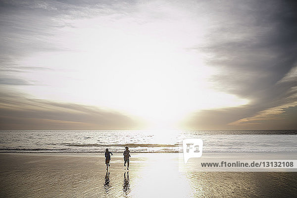 Verspielte Brüder rennen bei Sonnenuntergang am Strand gegen bewölkten Himmel