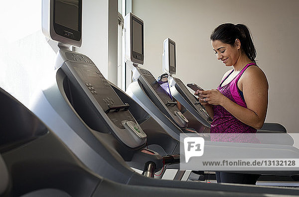 Lächelnde Frau benutzt Smartphone  während sie im Fitnessstudio auf dem Laufband steht