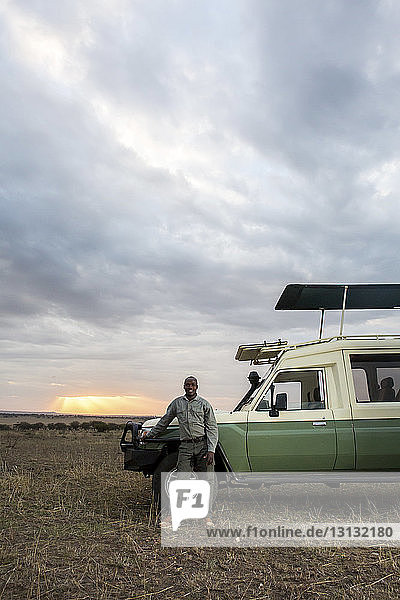 Porträt eines Mannes  der im Serengeti-Nationalpark vor bewölktem Himmel neben einem Geländewagen steht