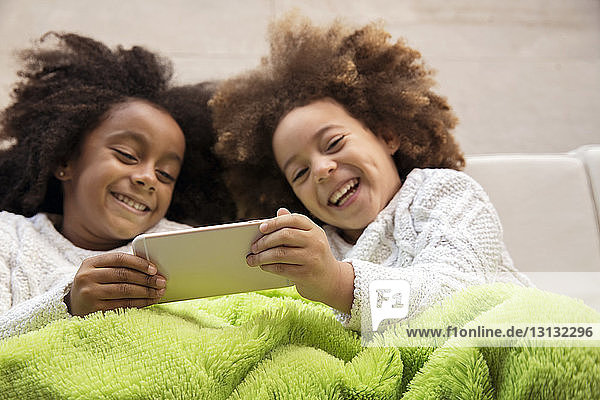Hochwinkelaufnahme von fröhlichen Schwestern  die auf dem Sofa liegen und ein Smartphone benutzen