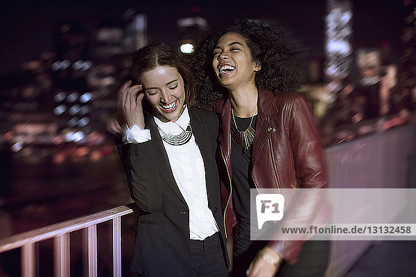 Female friends laughing while standing by railing on street at night