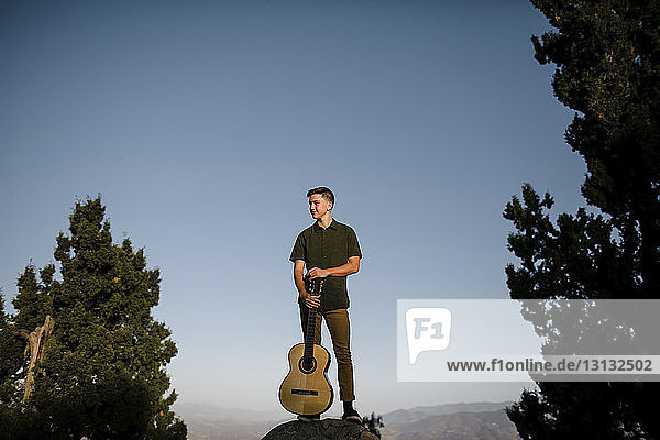 Mann hält Gitarre in der Hand  während er bei Sonnenuntergang auf einem Felsen vor klarem blauen Himmel steht
