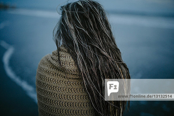 Rückansicht einer nassen Frau mit Schal, die in der Abenddämmerung am Strand steht
