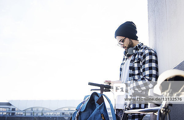 Young man using phone while leaning on wall by bicycle against clear sky
