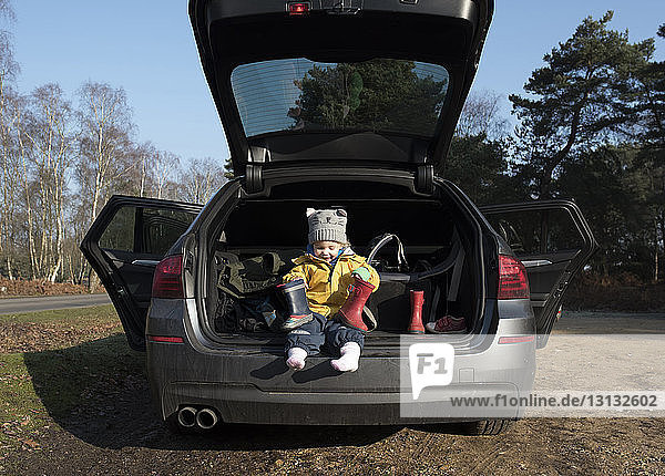 Süßes Mädchen hält Gummistiefel in der Hand  während es im Kofferraum eines Autos sitzt