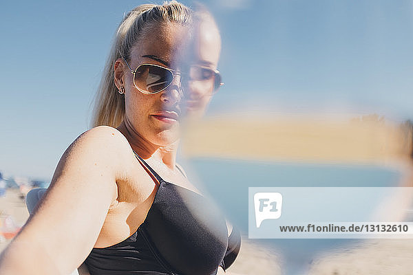 Porträt einer Frau mit Sonnenbrille und Bikini am Strand mit einem Trinkglas im Vordergrund