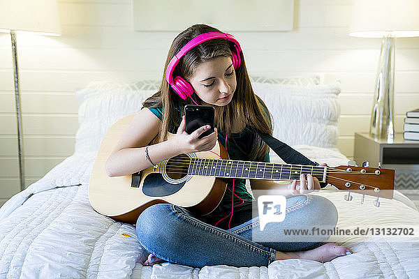 Teenager-Mädchen benutzt Smartphone beim Gitarrespielen im Schlafzimmer