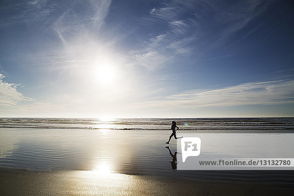 Silhouettenfrau joggt am sonnigen Tag am Meeresufer gegen den Himmel