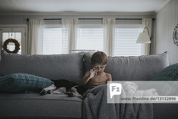Shirtless boy using tablet computer while sitting by dog on sofa at home