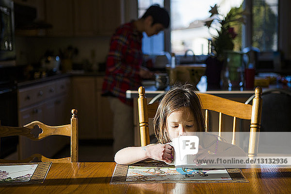 Tochter trinkt Tee auf dem Tisch  während der Vater zu Hause im Hintergrund arbeitet