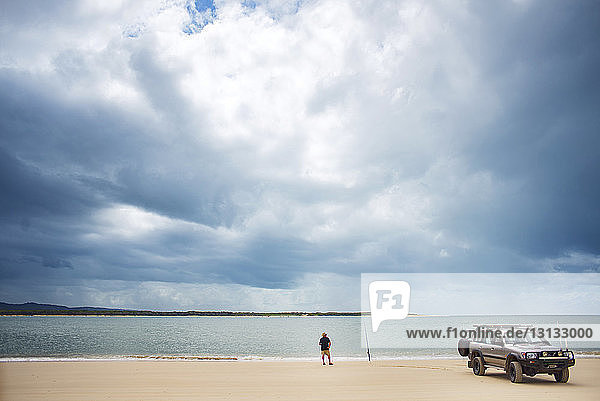 Rear view of senior man standing by off-road vehicle at Inskip Point against cloudy sky