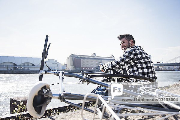 Young man sitting by bicycle in front of canal against clear sky