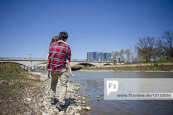 Full length of father carrying daughter while walking on riverbank against bridge and clear blue sky during sunny day