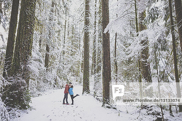 Full length of couple kissing while standing amidst trees in forest at Lynn Canyon Park during winter