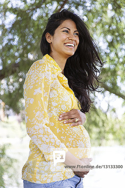 Glückliche schwangere Frau mit Händen auf dem Bauch steht im Park