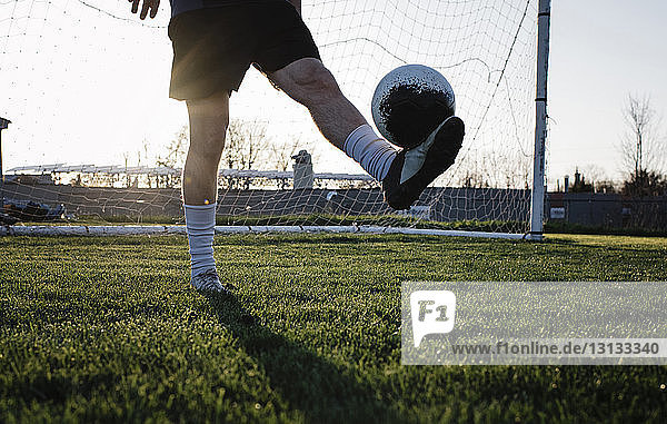 Low section of man practicing soccer on grassy field against clear sky during sunset