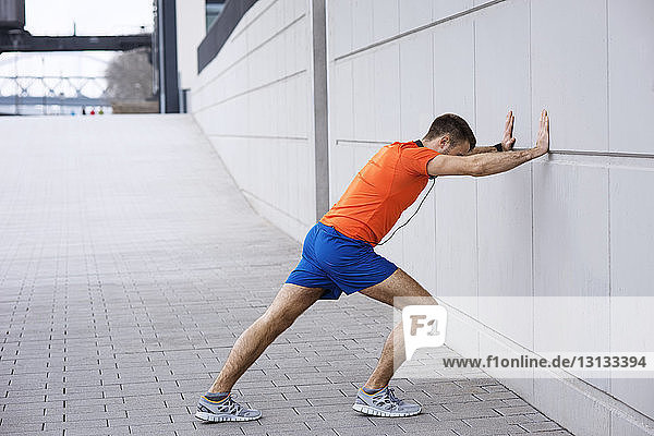 Side view of male athlete leaning on wall while exercising on footpath