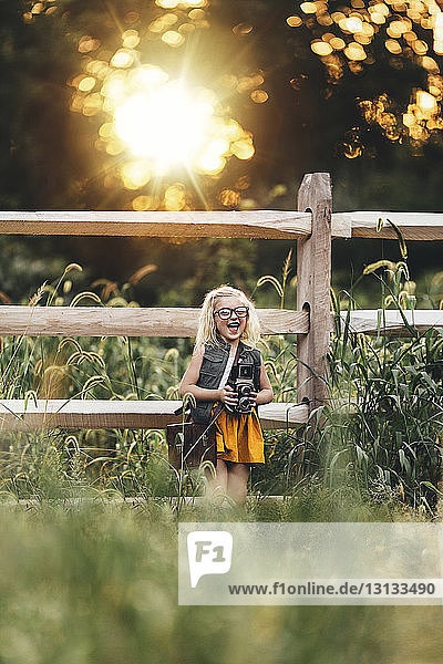 Cheerful girl with camera standing on grassy field against fence during sunset