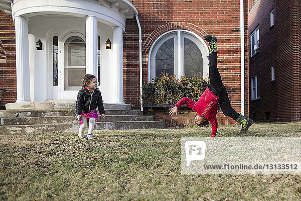 Sister looking at brother practicing cartwheel on lawn against house