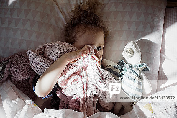 Overhead portrait of cute girl covering eye with textile while lying on bed at home