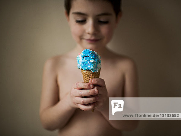 Shirtless boy holding ice cream while standing against wall at home
