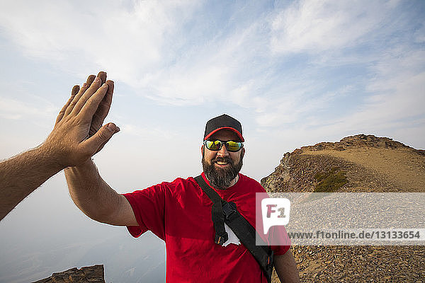 Cropped hand of man giving high-five to friend on mountains against cloudy sky