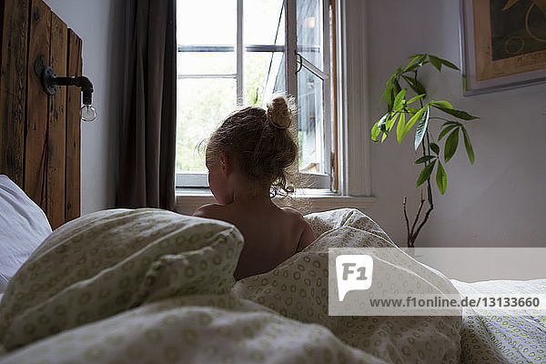 Rear view of shirtless girl sitting with duvet on bed at home