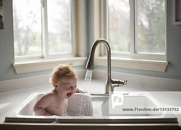 Cute shirtless baby boy sitting in kitchen sink against window at home