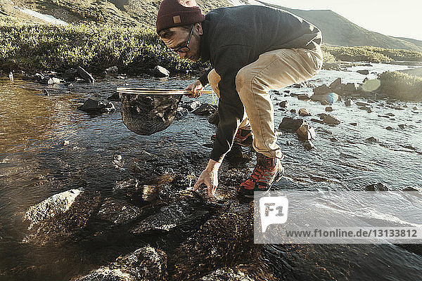 Mann hält Falterfischernetz in voller Länge  während er bei sonnigem Wetter Fische aus dem Fluss fängt