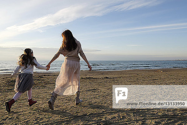 Rear view of mother and daughter holding hands while running at beach against sky during sunset