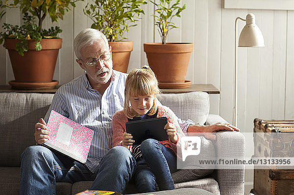 Girl using tablet computer while sitting with grandfather on sofa at home