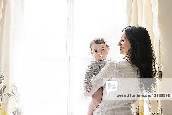 Porträt eines niedlichen Sohnes  den die Mutter zu Hause gegen das Fenster trägt