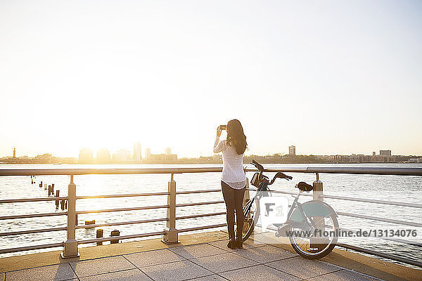 Rückansicht einer Frau  die die Stadt fotografiert  während sie bei Sonnenuntergang mit dem Fahrrad auf der Promenade steht