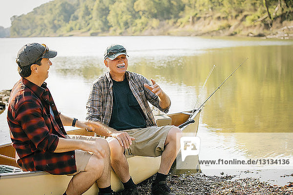 Happy male friends talking while sitting on boat at lakeshore