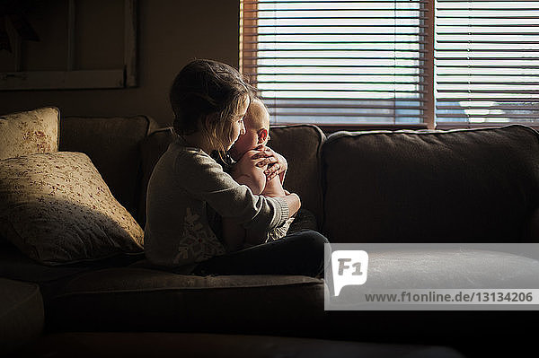 Seitenansicht eines Mädchens  das einen kleinen Jungen umarmt  während es zu Hause auf dem Sofa sitzt