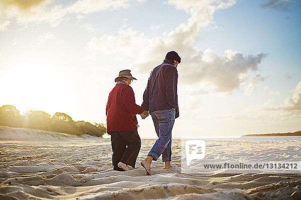 Senior couple holding hands while walking on sandy beach on sunny day