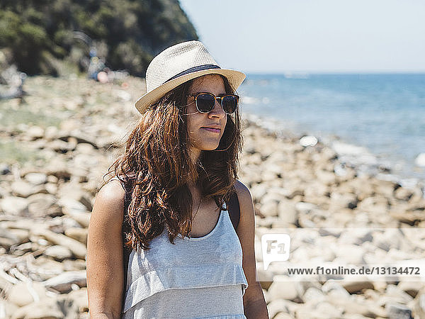 Woman wearing sunglasses and hat while standing at rocky beach against sky during sunny day