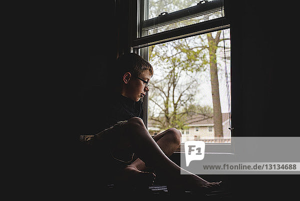 Nachdenklicher Junge schaut durchs Fenster  während er zu Hause in der Dunkelkammer sitzt