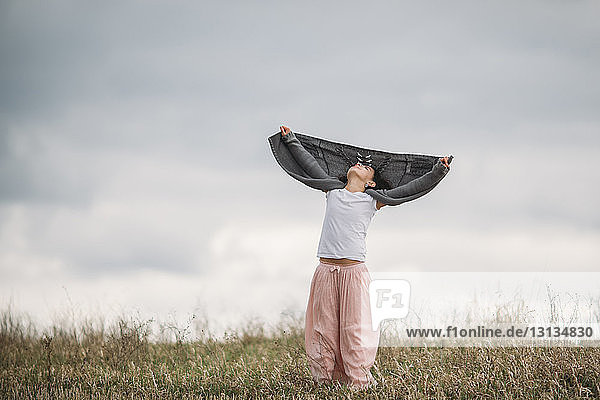 Mädchen hält Pullover in der Hand während sie auf dem Feld vor bewölktem Himmel im Park steht