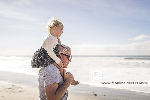 Side view of grandfather carrying granddaughter on shoulders while standing at beach against sky